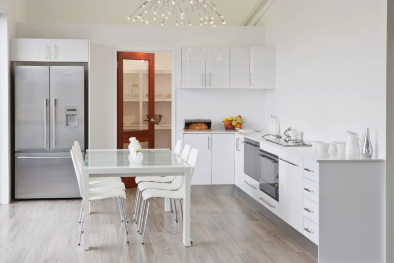 White kitchen area with metallic fridge, glass dining table and white dining furniture