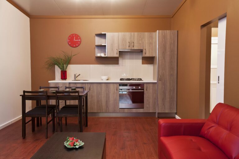 Open plan kitchen and living area with bright red couch, red carpet, wooden kitchen cabinets, stove, black wooden dining table and wooden coffee table