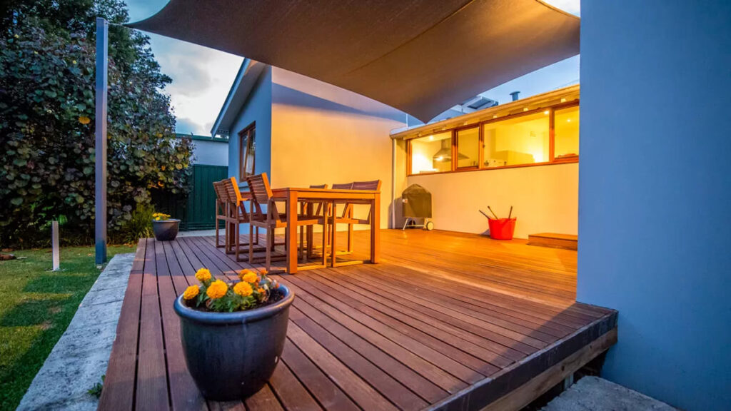 Backyard of house in the evening with deck, lawn, wooden outdoor furniture and a potted plant with orange flowers while house emanates light