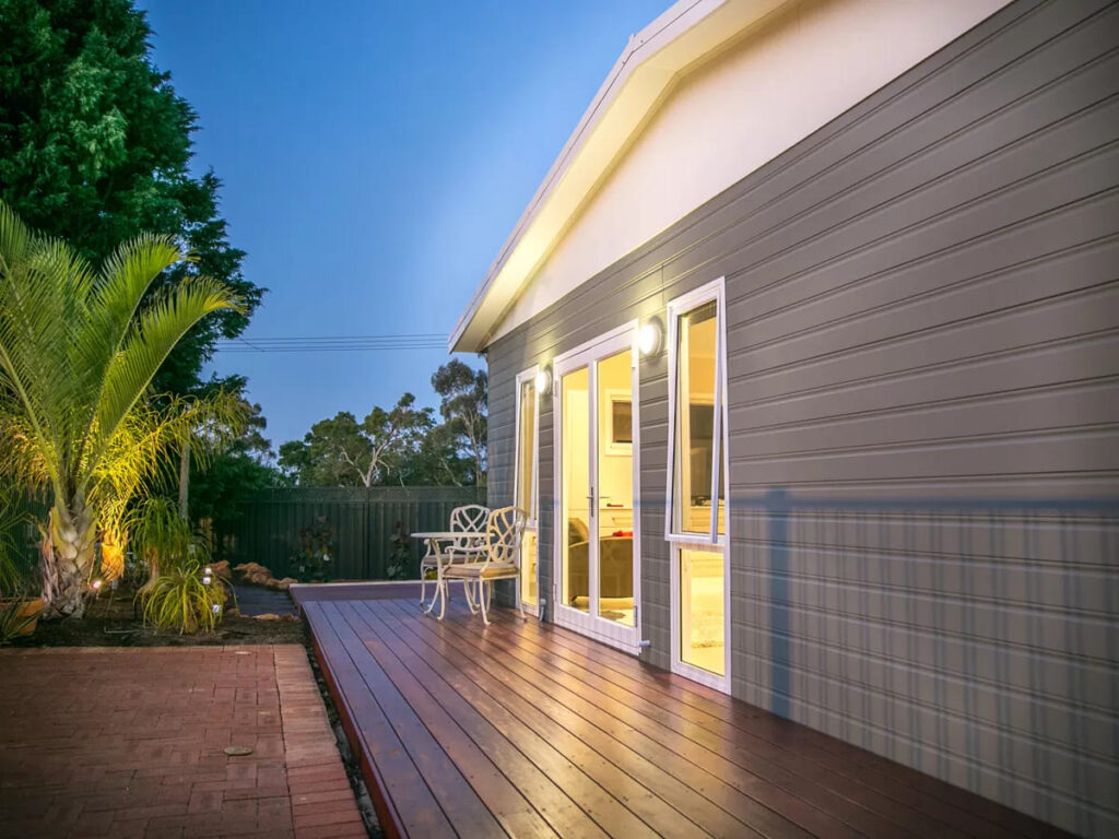 Backyard of grey granny flat in the evening emanating light onto wooden deck