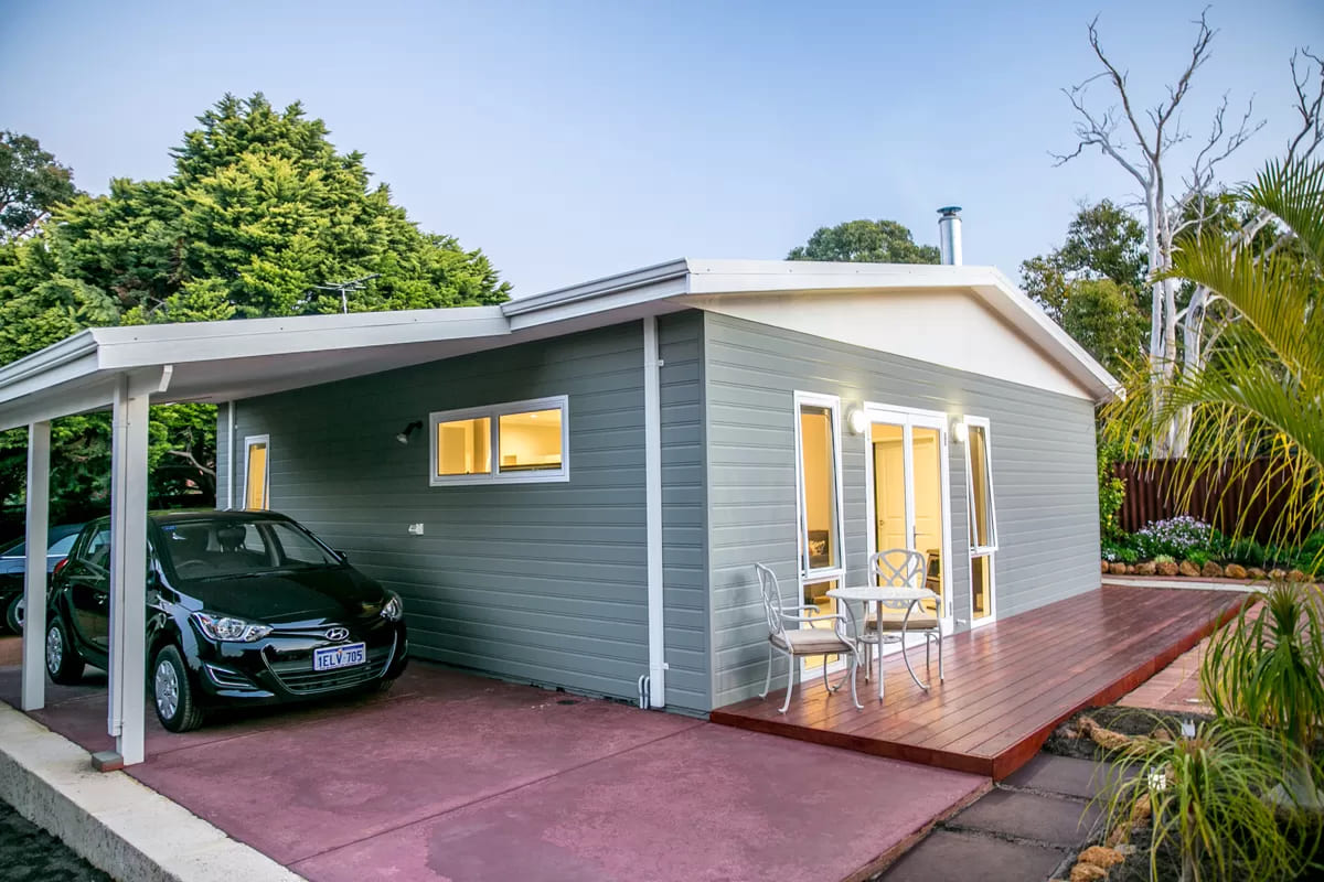 Exterior of grey granny flat with wooden deck, white outdoor furniture, sheltered driveway and black car parked