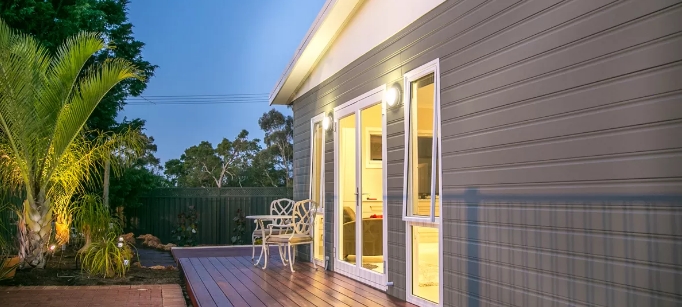 Wooden deck and exterior of grey granny flat. White alfresco dining chairs and light coming from inside the granny flat.