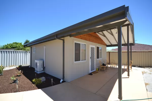 White granny flat with awning, outdoor furniture and garden area with potted plants. 