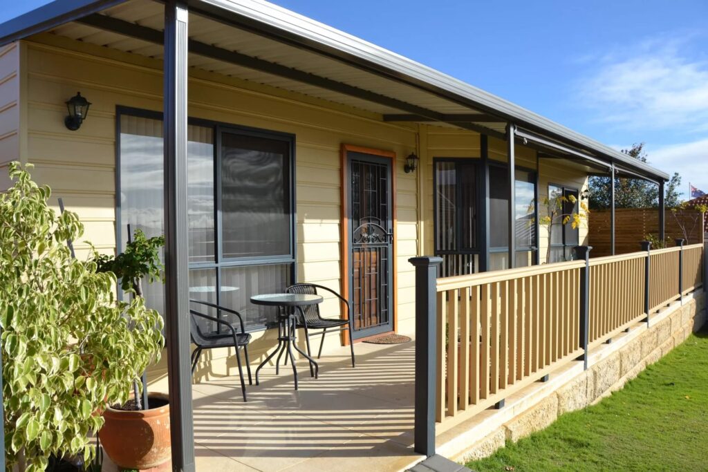 Front of yellow house with deck, black alfresco furniture, wooden fence and potted plants on a sunny day. 